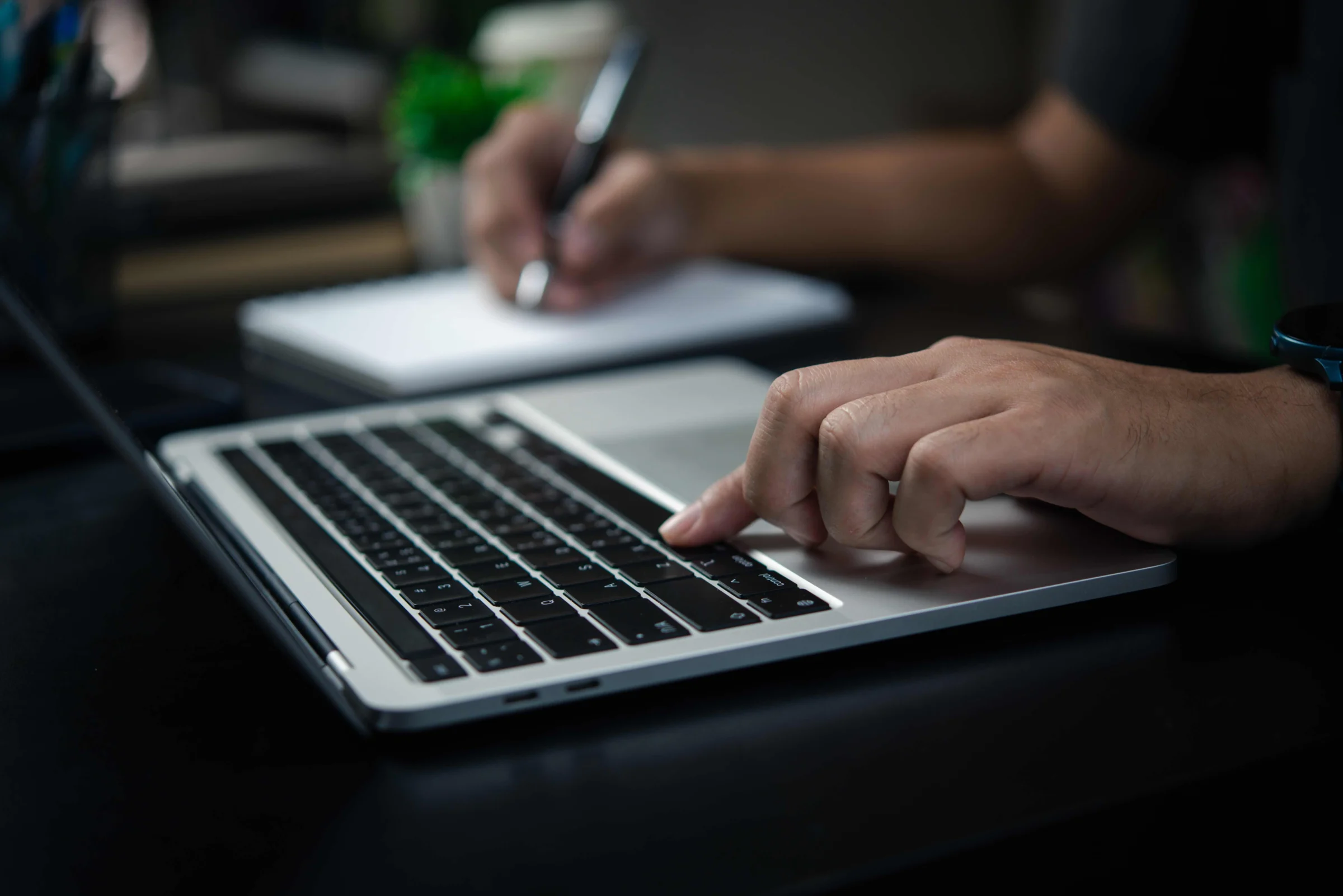 A filmmaker typing on a laptop at night while making handwritten notes on a notepad beside it